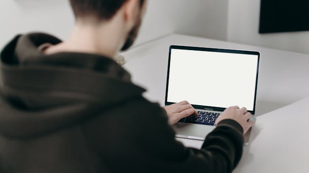 A young man in a hoodie working on a laptop with a blank screen at a minimal home office desk.